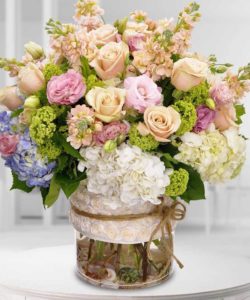 Premium floral varieties of roses, hydrangea, greens and more are filled in a glass vase bearing a designer ribbon.