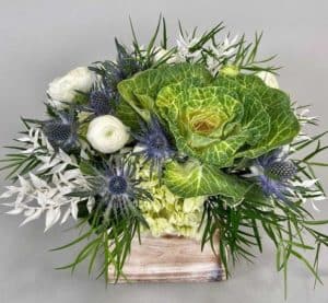 Mix of greenery, green hydrangea, kale, white dendrobium orchids, blue delphium, cream stock arranged in a blue ceramic vase